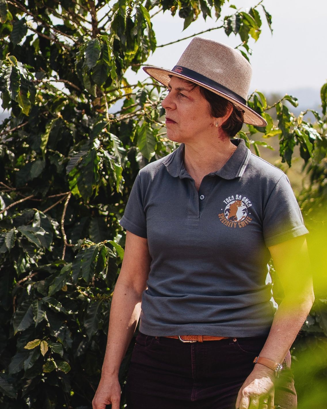 Luciana Flores Martins Swan in a Toca da Onça shirt and straw hat, standing beside a coffee tree